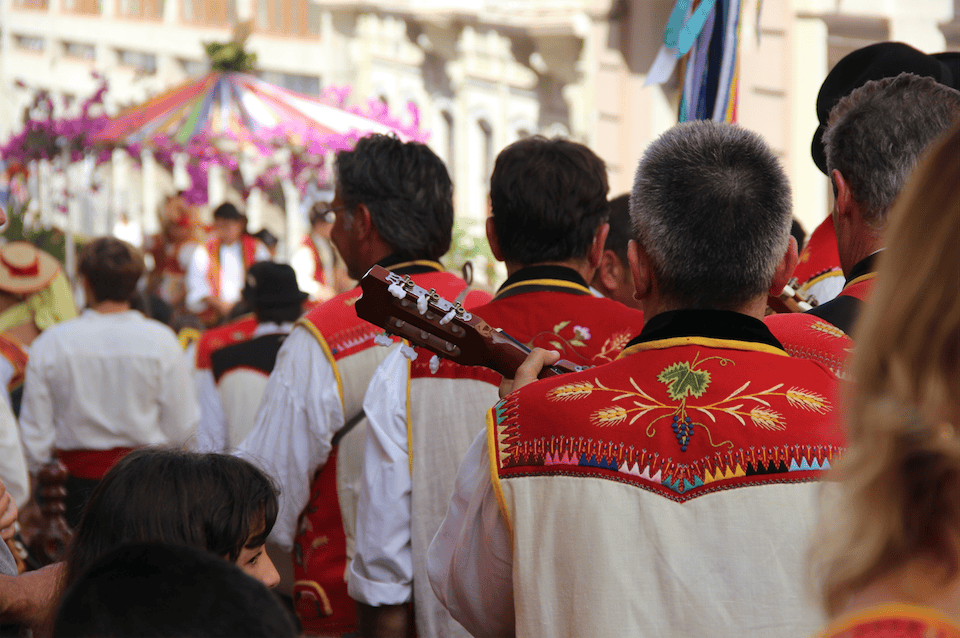 Grupo de personas con vestimenta canaria típica y tradicional durante una romería, con chalecos bordados en rojo y blanco mientras avanzan entre música y ambiente festivo.